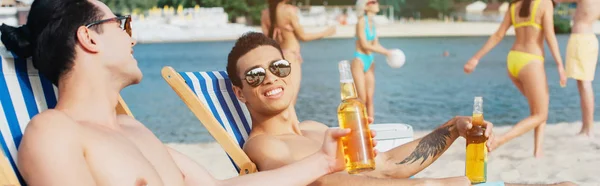 Panoramic shot of multicultural friends sitting in chaise lounges and holding bottles with beer — Stock Photo