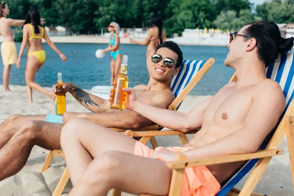 Amigos multiculturais bonitos conversando enquanto sentados na praia em salões de chaise com garrafas de cerveja — Fotografia de Stock