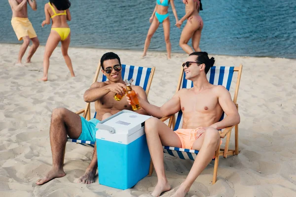 Cheerful multicultural friends clinking bottles of beer while sitting in chaise lounges near portable fridge on beach — Stock Photo