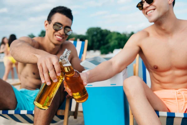 Amigos multiculturales sonrientes tintineando botellas de cerveza mientras están sentados en chaise lounges en la playa - foto de stock