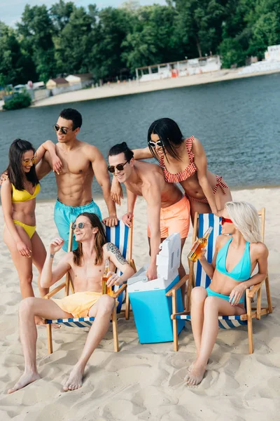 Allegro uomo e donna seduti in chaise longue con bottiglie di birra vicino agli amici multiculturali sulla spiaggia — Foto stock