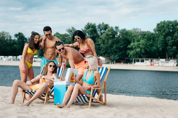 Cheerful multicultural friends standing near man and woman sitting in chaise lounges with bottles of beer — Stock Photo