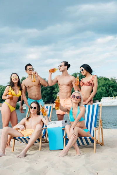 Jóvenes amigos multiculturales bebiendo cerveza mientras descansan en la playa - foto de stock