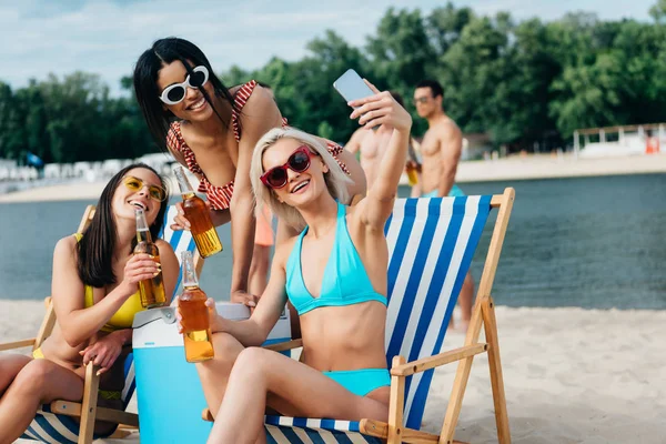 Hermosas chicas multiculturales en trajes de baño tomando selfie mientras bebe cerveza en la playa - foto de stock