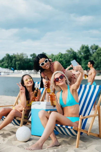 Meninas multiculturais bonitas em maiôs tomando selfie enquanto bebe cerveja na praia — Fotografia de Stock