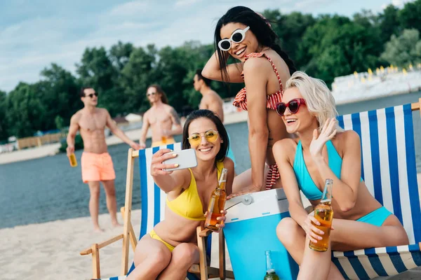 Hermosas chicas multiculturales tomando selfie mientras se divierten en la playa cerca de hombres jóvenes guapos - foto de stock