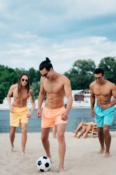 Homens multiculturais bonitos e sem camisa a jogar futebol na praia — Fotografia de Stock