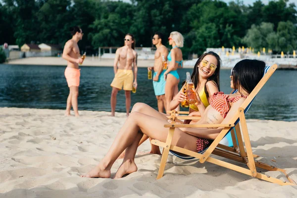 Meninas multiculturais alegres bebendo cerveja em salões de chaise perto de amigos descansando na praia — Fotografia de Stock