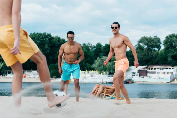 Gutaussehende, hemdlose multikulturelle Freunde, die am Strand Fußball spielen — Stockfoto