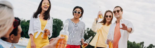 Panoramic shot of cheerful multicultural friends with bottles of beer having fun on beach — Stock Photo
