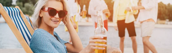 Панорамный снимок молодой женщины в солнечных очках, улыбающейся в камеру, сидящей в шезлонге с бутылкой пива — Stock Photo