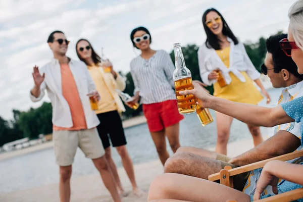 Smiling multicultural friends in sunglasses drinking beer while resting on beach — Stock Photo