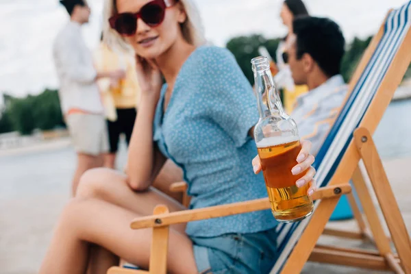 Enfoque selectivo de mujer joven atractiva en gafas de sol sosteniendo botella de cerveza mientras está sentado en el chaise lounge cerca de amigos multiculturales - foto de stock