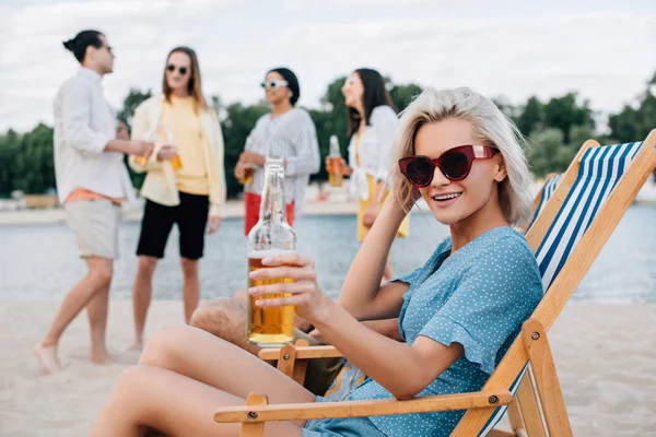 Веселая молодая женщина улыбается в камеру, сидя в шезлонге с бутылкой пива рядом с мультикультурными друзьями — стоковое фото