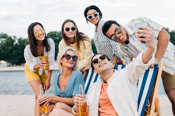 Schöner junger Mann macht Selfie mit multikulturellen Freunden, die Spaß haben und Bier am Strand trinken — Stockfoto