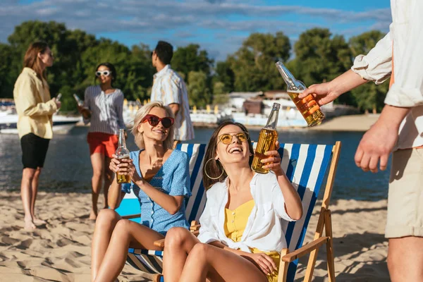 Jóvenes, alegres hombres y mujeres multiculturales bebiendo cerveza mientras descansan en la playa - foto de stock