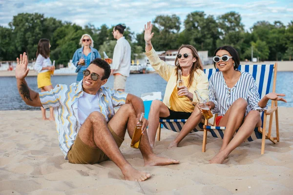Handsome multicultural men waving hands while having fun with friends on beach — Stock Photo