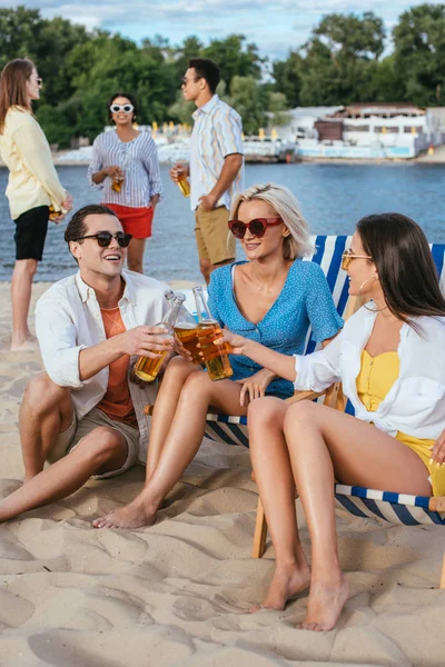 Beau jeune homme cliquetis bouteilles de bière avec des filles gaies tout en s'amusant sur la plage avec des amis multiculturels — Photo de stock
