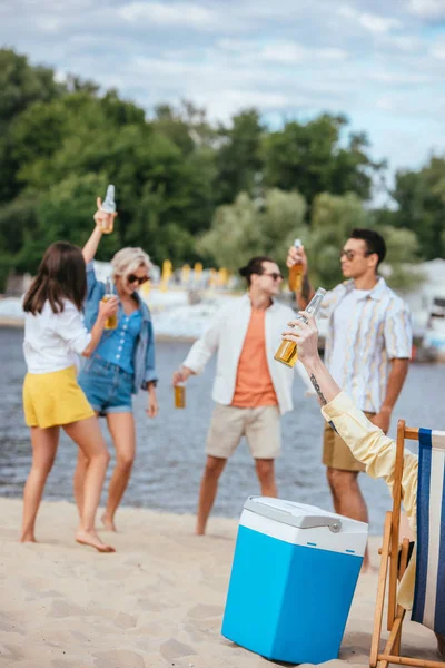 Избирательный фокус веселых мультикультурных друзей, держащих бутылки пива, веселясь на пляже — стоковое фото