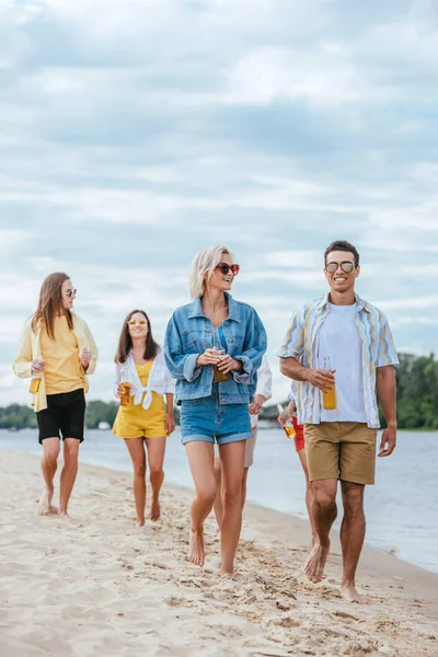 Happy multicultural friends walking on riverside and holding bottles of beer — Stock Photo