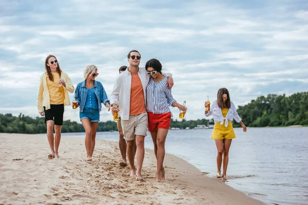 Pareja interracial abrazándose mientras camina a orillas del río junto con amigos multiculturales - foto de stock