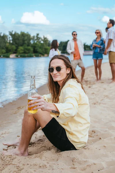 Sonriente joven mirando a la cámara mientras está sentado en la playa y sosteniendo la botella de cerveza - foto de stock