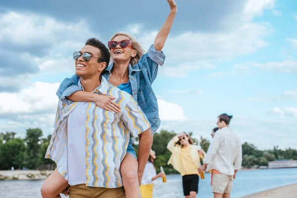 Guapo bi-racial hombre piggybacking novia mientras camina en la playa cerca de amigos multiculturales - foto de stock