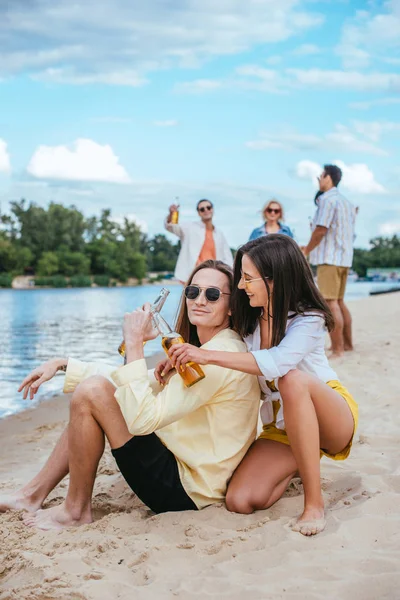 Feliz joven pareja tintineo botellas de cerveza mientras está sentado en la playa cerca de amigos multiculturales - foto de stock