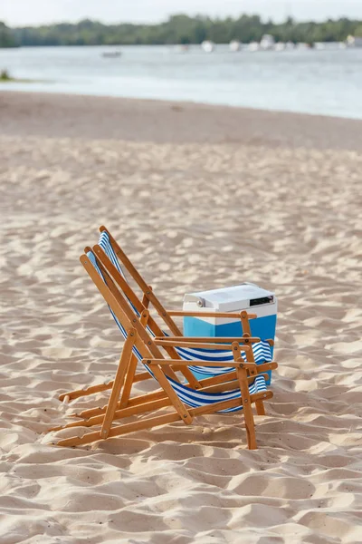 Dos chaise lounges y nevera portátil en la playa de arena cerca del río - foto de stock