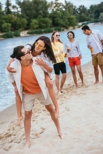 Beau jeune homme piggyback heureux copine tout en s'amusant sur la plage avec des amis multiculturels — Photo de stock