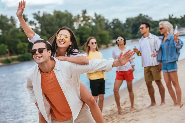 Весёлый молодой человек, подкатывающий к счастливой девушке, гуляя по пляжу вместе с мультикультурными друзьями — стоковое фото