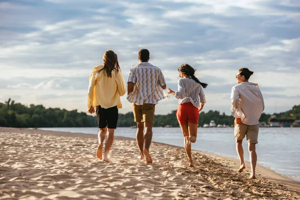 Vista trasera de amigos multiculturales corriendo en la playa cerca del río - foto de stock