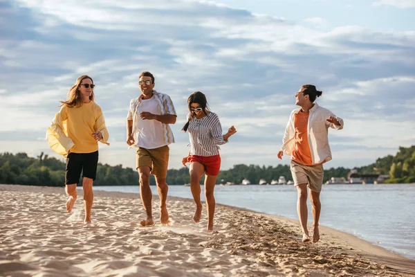 Alegres amigos multiculturales corriendo en la playa de arena cerca del río - foto de stock