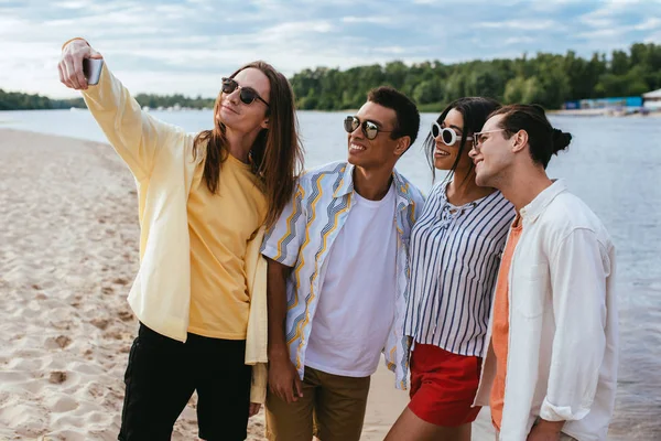 Sorridente giovane che prende selfie con allegri amici multiculturali sulla spiaggia — Foto stock