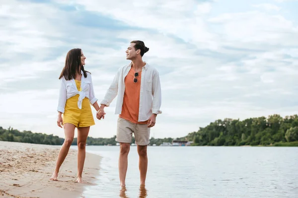 Feliz pareja joven cogidos de la mano y mirándose mientras caminan a orillas del río - foto de stock