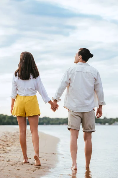 Back view of young couple holding hands and looking at each other while walking on riverside — Stock Photo