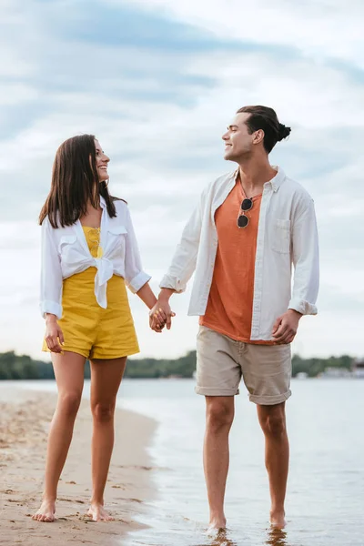 Joyeux jeune couple tenant la main et se regardant tout en marchant sur la plage — Photo de stock