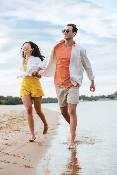 Feliz joven pareja cogida de la mano mientras se ejecuta en la playa cerca del río - foto de stock