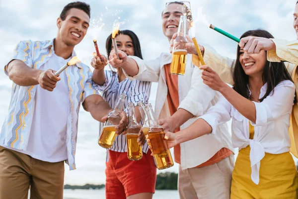 Amigos multiculturales felices sosteniendo chispas y botellas de cerveza mientras se divierten en la playa - foto de stock