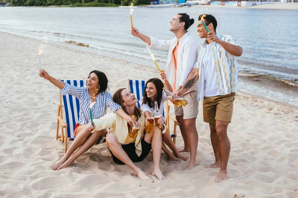 Cheerful multicultural friends holding bottles of beer and sparklers while having fun on beach — Stock Photo