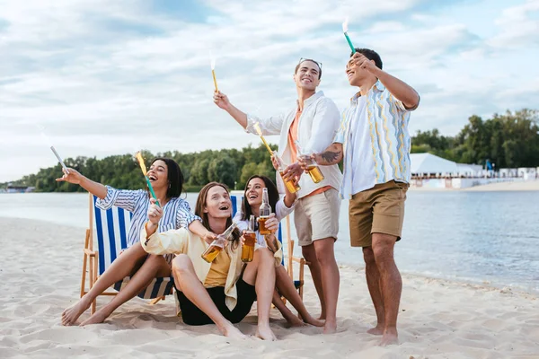 Happy multicultural friends having fun on beach while holding sparklers and bottles of beer — Stock Photo