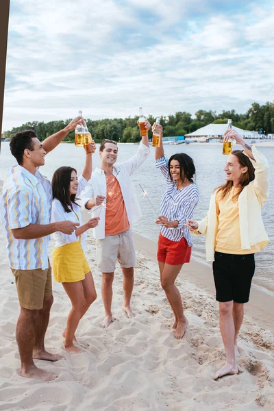 Amigos multiculturales sonrientes sosteniendo botellas de cerveza y bengalas mientras se divierten en la playa - foto de stock