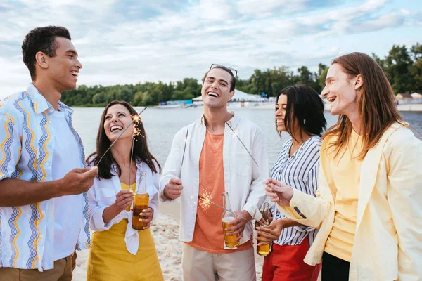 Heureux amis multiculturels tenant des bouteilles de bière et des étincelles tout en s'amusant sur la plage — Photo de stock