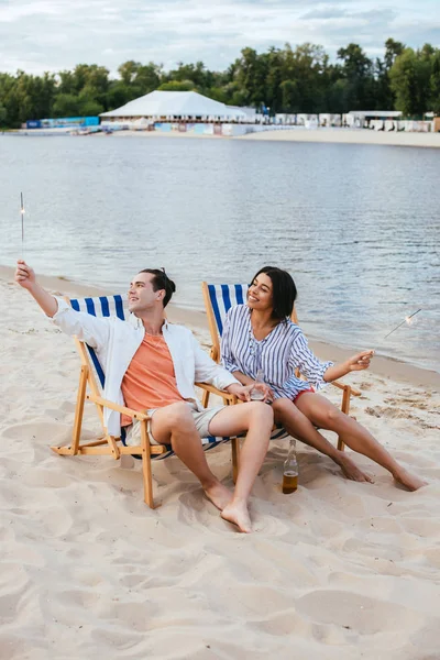 Heureux couple interracial assis dans des chaises longues et tenant des étincelles tout en se reposant sur la plage — Photo de stock