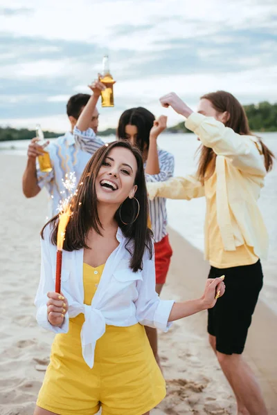 Heureuse jeune femme tenant scintillant et regardant la caméra tout en s'amusant sur la plage près d'amis multiculturels — Photo de stock