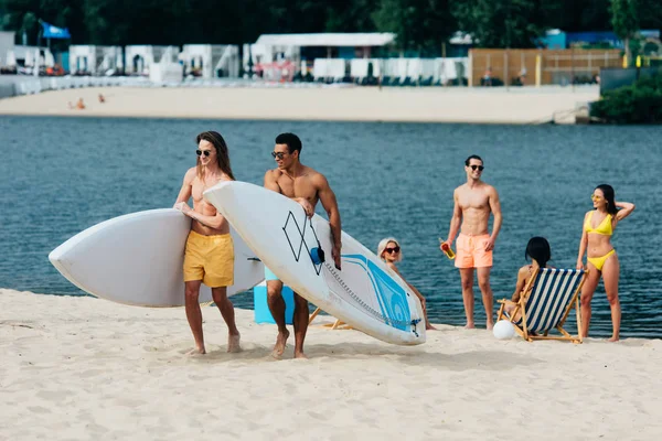 Guapo, jóvenes hombres multiculturales sosteniendo tablas de surf mientras caminan en la playa - foto de stock