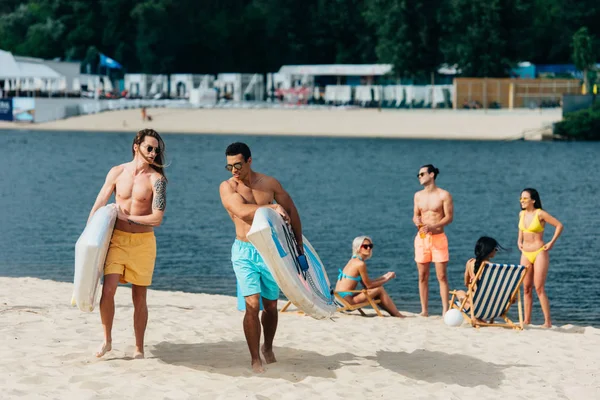 Homens multiculturais bonitos andando na praia e segurando pranchas de surf — Fotografia de Stock