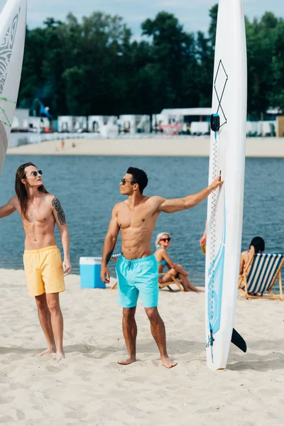 Gutaussehende, junge multikulturelle Männer stehen am Strand in der Nähe von Surfbrettern — Stockfoto