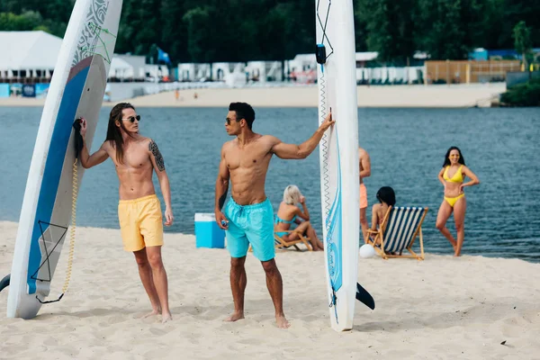 Handsome, young multicultural friends standing on beach near surfing boards — Stock Photo