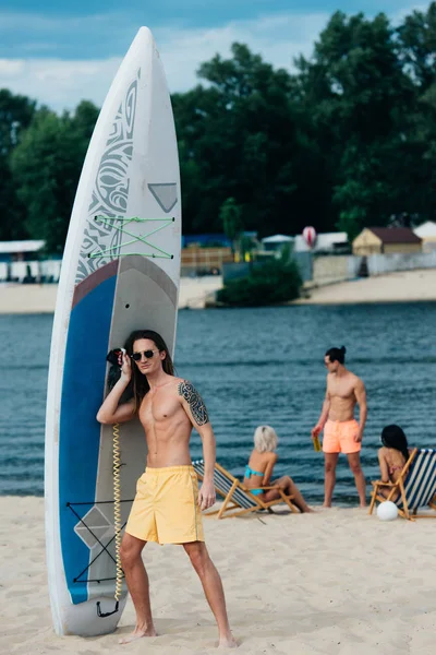 Jovem bonito olhando de pé perto da prancha de surf na praia e olhando para a câmera — Fotografia de Stock